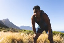 Hombre afroamericano haciendo ejercicio al aire libre descansando en una montaña. entrenamiento de fitness y estilo de vida saludable al aire libre. - foto de stock