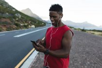 Hombre afroamericano haciendo ejercicio al aire libre usando un smartphone en una carretera costera. entrenamiento de fitness y estilo de vida saludable al aire libre. - foto de stock