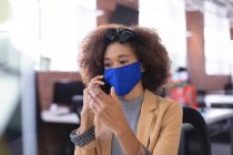 Femme d'affaires afro-américaine portant un masque parlant par smartphone au bureau. entreprise indépendante de conception créative pendant la pandémie de coronavirus covid 19. — Photo de stock