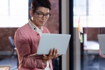 Стильный азиатский бизнесмен, стоящий и пользующийся ноутбуком. деловой человек за работой в современном офисе. — стоковое фото