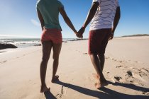 Mittelteil eines afrikanisch-amerikanischen Paares, das Händchen haltend am Strand spaziert. gesunde Freizeit im Freien am Meer. — Stockfoto