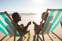 Pareja afroamericana enamorada sentada en tumbonas, disfrutando de bebidas en la playa. amor, romance y vacaciones de verano de vacaciones de playa. - foto de stock