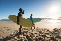 Feliz pareja afroamericana en la playa llevando tablas de surf. tiempo de ocio al aire libre saludable junto al mar. - foto de stock