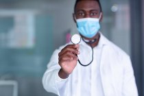 Retrato de um médico afro-americano vestindo máscara facial segurando estetoscópio na câmera. profissional médico a trabalhar durante a pandemia do coronavírus covid 19. — Fotografia de Stock