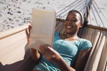 Mujer afroamericana feliz acostada en hamaca en la playa usando tableta. tiempo de ocio al aire libre saludable junto al mar. - foto de stock