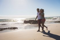 Счастливая африканская американская пара, гуляющая по пляжу обнимаясь. здоровый отдых на открытом воздухе у моря. — стоковое фото