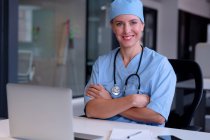 Retrato de uma médica caucasiana sorridente sentada à secretária a usar esfoliantes usando um computador portátil. profissional médico no trabalho. — Fotografia de Stock