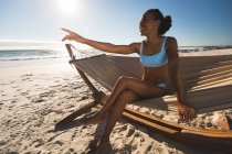 Happy african american woman sitting in hammock on beach pointing. healthy outdoor leisure time by the sea. — Stock Photo