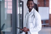 Médecin afro-américain souriant portant un manteau blanc et un stéthoscope à l'aide d'une tablette numérique. professionnel de la santé au travail. — Photo de stock