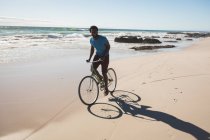 Happy african american man on beach riding bicycle. healthy outdoor leisure time by the sea. — Stock Photo