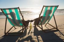 African american couple in love sitting in deckchairs, holding hands on beach. love, romance and beach break summer holiday. — Stock Photo