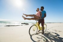Feliz pareja afroamericana montando bicicletas en la playa. tiempo de ocio al aire libre saludable junto al mar. - foto de stock