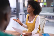 Feliz mujer de negocios afroamericana sosteniendo documento hablando con colegas en la sala de reuniones. negocio independiente de diseño creativo. - foto de stock