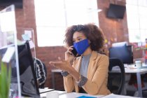 Африканская американка в маске разговаривает по смартфону в офисе. независимый бизнес креативного дизайна во время пандемии коронавируса ковида 19. — стоковое фото