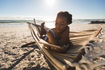 Mujer afroamericana feliz acostada en hamaca en la playa usando tableta. tiempo de ocio al aire libre saludable junto al mar. - foto de stock