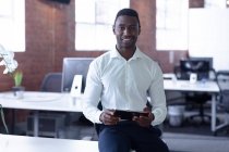 Retrato de sonriente hombre de negocios afroamericano casual sentado en el escritorio con la tableta. persona de negocios en el trabajo en la oficina moderna. - foto de stock