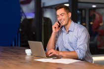 Retrato de homem de negócios caucasiano casual feliz falando no smartphone na mesa usando laptop. pessoa de negócios no trabalho em escritório moderno. — Fotografia de Stock