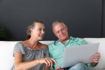 Feliz casal sênior caucasiano sentado na sala de estar olhando para laptop falando e sorrindo. ficar em casa em isolamento durante o confinamento de quarentena. — Fotografia de Stock