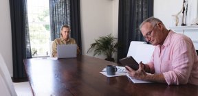 Couple âgé caucasien dans le salon assis à la table de travail, en utilisant un ordinateur portable et une tablette. rester à la maison dans l'isolement pendant le confinement en quarantaine. — Photo de stock