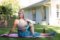 Старшая белая женщина, занимающаяся спортом в саду, сидящая на коврике для йоги. оставаться дома в изоляции во время карантинной изоляции. — стоковое фото