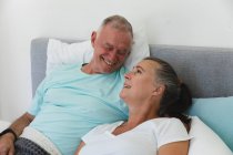 Счастливая старшая кавказская пара отдыхает в спальне, сидит в постели и улыбается друг другу. оставаться дома в изоляции во время карантинной изоляции. — стоковое фото