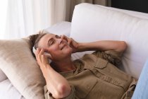 Mujer mayor caucásica feliz en la sala de estar acostado en el sofá con auriculares y sonriendo. permanecer en casa aislado durante el bloqueo de cuarentena. - foto de stock
