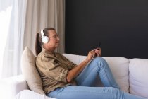 Mulher idosa caucasiana feliz na sala de estar sentada no sofá usando fones de ouvido e usando smartphone. ficar em casa em isolamento durante o confinamento de quarentena. — Fotografia de Stock