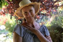 Retrato de mulher sênior caucasiana feliz em pé no jardim ensolarado usando chapéu de sol, sorrindo para a câmera. ficar em casa em isolamento durante o confinamento de quarentena. — Fotografia de Stock
