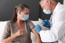 Caucasian senior male doctor giving female patient covid 19 vaccination, both wearing face masks. medical professional at work during coronavirus covid 19 pandemic. — Stock Photo