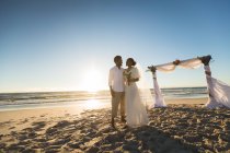 Счастливая африканская американская пара влюблённая выходит замуж, обнимается на пляже во время заката. любовь, романтика и свадебный пляжный отдых. — стоковое фото