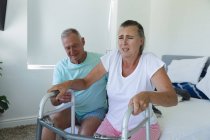 Caucasian senior couple sitting on bed, husband helping wife stand using walking frame. staying at home in isolation during quarantine lockdown. — Stock Photo