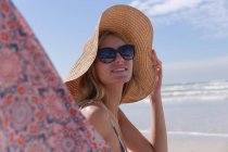Улыбающаяся белая женщина в бикини и шляпе, сидящая на шезлонге и смотрящая в камеру на пляже. здоровый отдых на открытом воздухе у моря. — стоковое фото