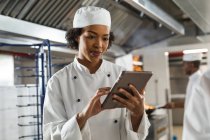 Портрет змішаної раси жіночого професійного шеф-кухаря, який використовує планшет з колегою на задньому плані. робота в зайнятій кухні ресторану . — стокове фото