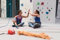 Two caucasian women talking and giving high five sitting on floor at indoor climbing wall. fitness and leisure time at gym. — Stock Photo