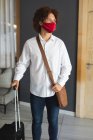 Portrait of mixed race man wearing face mask standing in hotel lobby with shoulder bag and suitcase. business travel hotel during coronavirus covid 19 pandemic. — Stock Photo