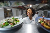 Gemischte Rasse professionelle Koch verlosen Gerichte, die mit Kollegen im Hintergrund serviert werden. Arbeit in einer belebten Restaurantküche. — Stockfoto