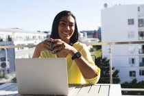 Усміхнена змішана транссексуальна жінка сидить за столом на сонячній терасі, використовуючи ноутбук, що тримає каву. перебування вдома в ізоляції під час карантину . — стокове фото