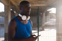 Afro-americano exercitando ao ar livre, usando fones de ouvido, usando smartphone sob ponte. treinamento de fitness ao ar livre saudável. — Fotografia de Stock