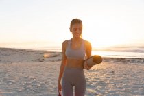 Donna caucasica che tiene il tappetino da yoga sorridente mentre sta in piedi sulla spiaggia. fitness yoga e stile di vita sano concetto — Foto stock