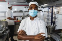 Портрет африканского американского профессионального шеф-повара в маске с коллегами на заднем плане. работа в оживленном ресторане кухни во время коронавируса ковид 19 пандемии. — стоковое фото