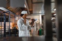 Portrait de cuisinière professionnelle métissée utilisant une tablette avec un collègue en arrière-plan. travailler dans une cuisine de restaurant occupée. — Photo de stock