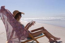 Mujer caucásica con bikini sentado en la silla de cubierta libro de lectura en la playa. tiempo de ocio al aire libre saludable junto al mar. - foto de stock