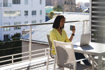 Смешанная расовая трансгендерная женщина, сидящая за столом на солнечной террасе на крыше, пьет кофе. оставаться дома в изоляции во время карантинной изоляции. — стоковое фото