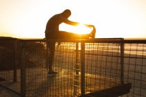 Hombre afroamericano haciendo ejercicio al aire libre, estirándose en el puente al atardecer. estilo de vida al aire libre saludable entrenamiento fitness. - foto de stock