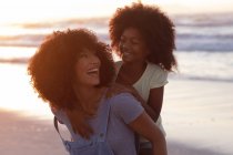 Smiling african american mother carrying her daughter piggyback at the beach smiling. healthy outdoor leisure time by the sea. — Photo de stock