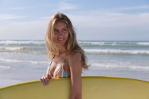 Smiling caucasian woman wearing bikini carrying yellow surfboard at the beach. healthy outdoor leisure time by the sea. - foto de stock
