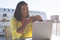 Sorridente donna transgender mista seduta a tavola sulla terrazza soleggiata con computer portatile in possesso di caffè. stare a casa in isolamento durante la quarantena. — Foto stock