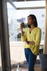 Smiling mixed race transgender woman standing on sunny roof terrace holding coffee. staying at home in isolation during quarantine lockdown. — Stock Photo