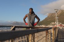 African american man exercising outdoors, resting on pier at sunset. healthy outdoor lifestyle fitness training. — Stock Photo