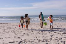 African american parents and two children holding beach accessories walking at the beach. family outdoor leisure time by the sea during coronavirus covid 19 pandemic. — Stock Photo